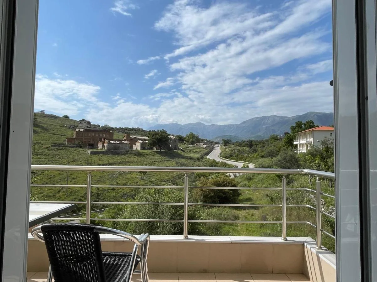 A balcony with a steel rail looking across scrub and trees to a derelict brick complex on the slope and the mountain ridge behind it