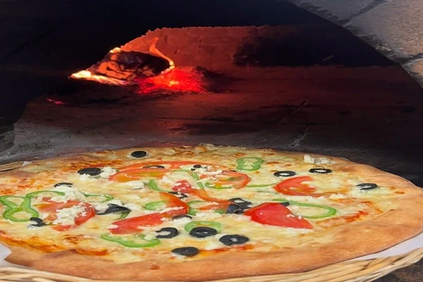 A pizza on a wooden peel in front of the lit wood-fired oven