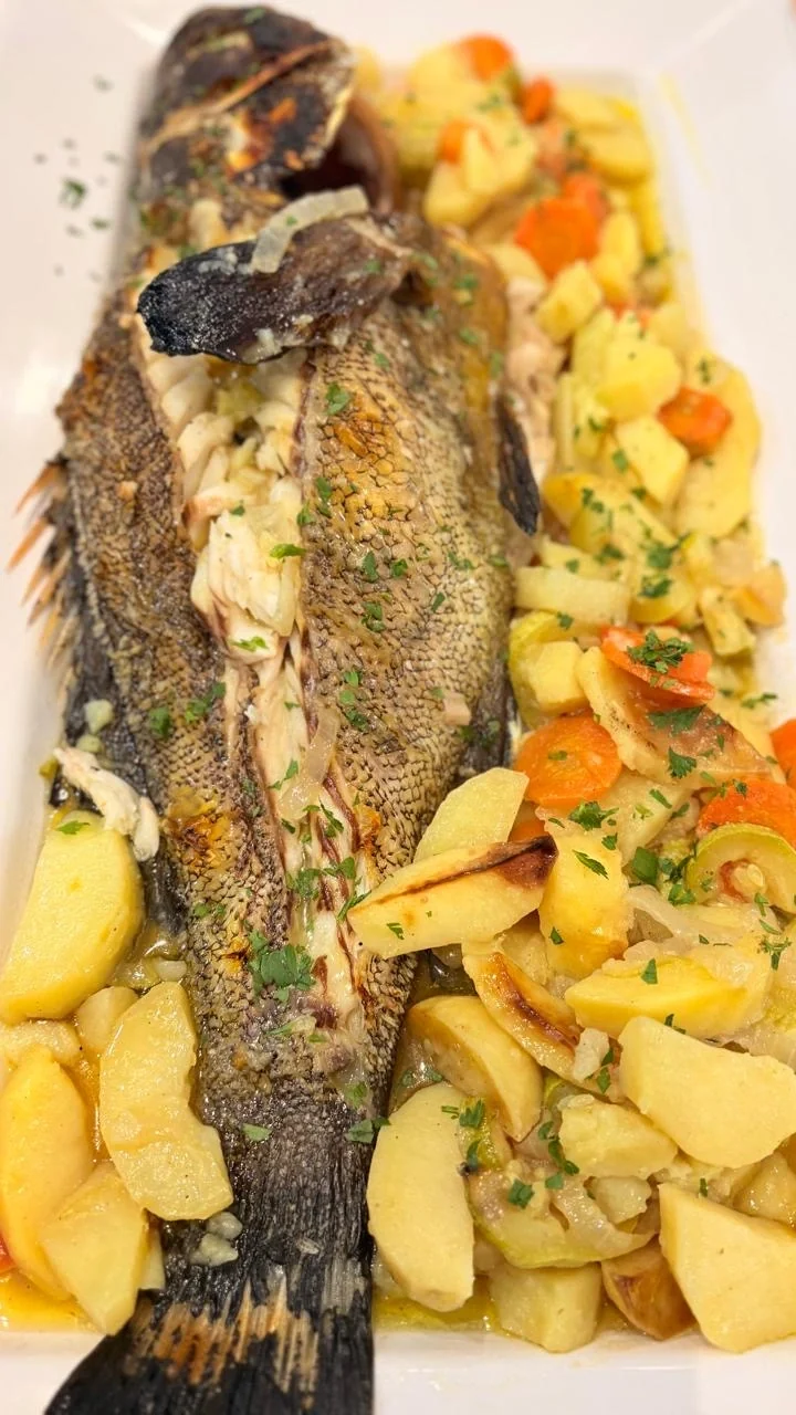 A whole baked fish on a white platter with potatoes, carrots, courgette and parsley