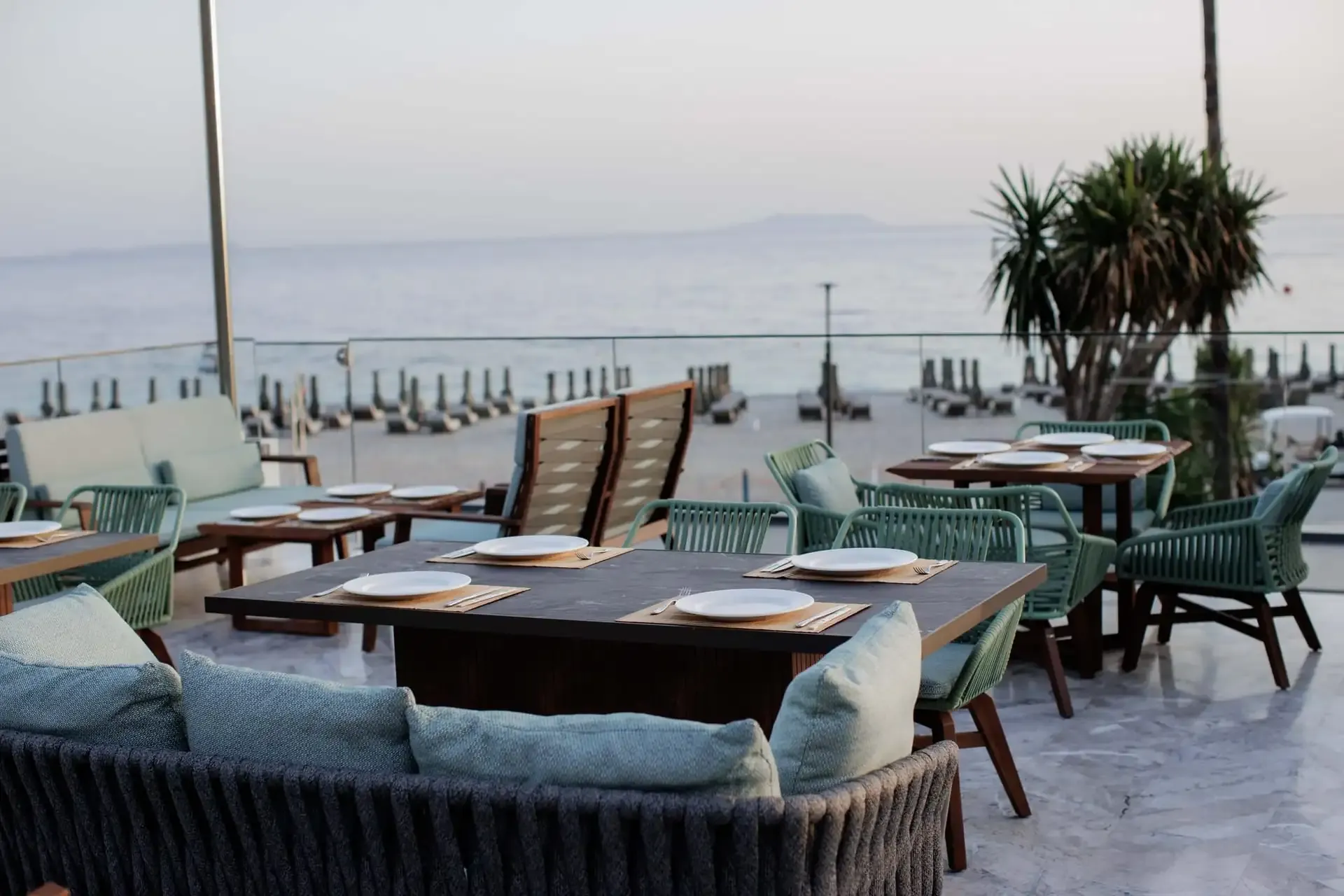 A sea-facing dining terrace at Prado, green rope chairs and laid slate tables above the beach and its rows of umbrellas