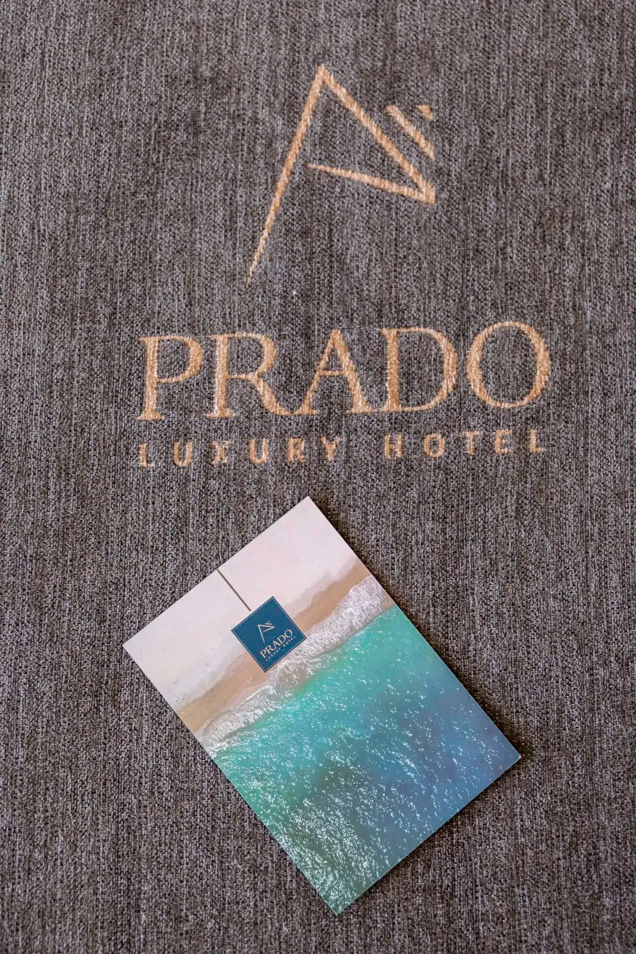 The Prado sail mark and wordmark stamped in gold foil into brown woven cloth, a printed card resting on it