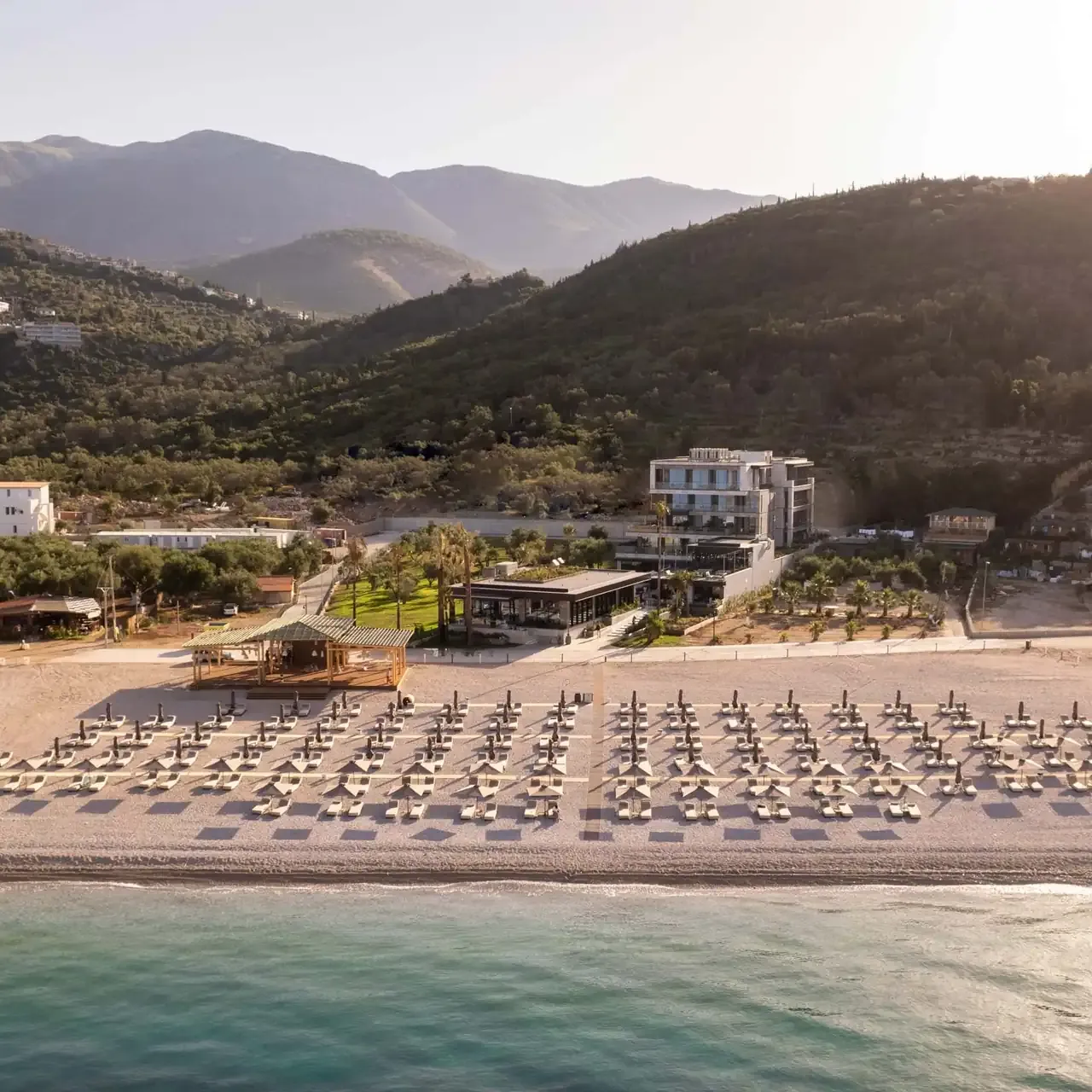 Prado Luxury Hotel seen from the water: the building low against a green olive slope, mountains behind it, and the ruled grid of its umbrellas on the shingle in front