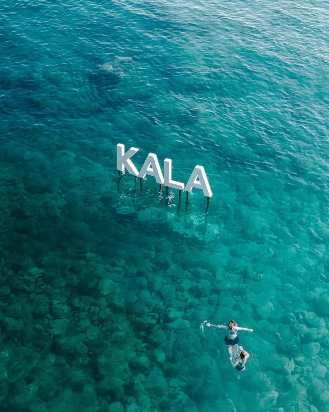 Aerial view of the iconic KALA letters standing in the turquoise shallows off Dhermi beach, with swimmers floating nearby — the Albanian Riviera setting of Kala Festival