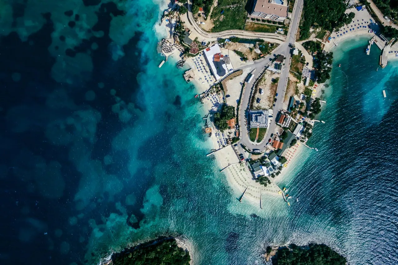 Aerial of the Ksamil promontory and its beach clubs — the busiest stretch of the destination most buses from Tirana drop near