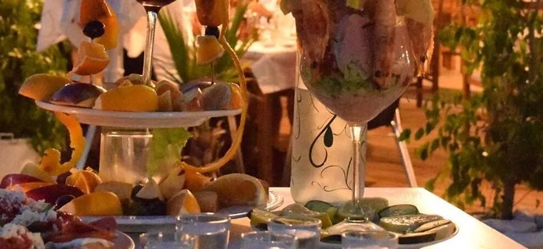 Prawns in a stemmed glass beside a tiered stand of citrus and shellfish on a laid table after dark