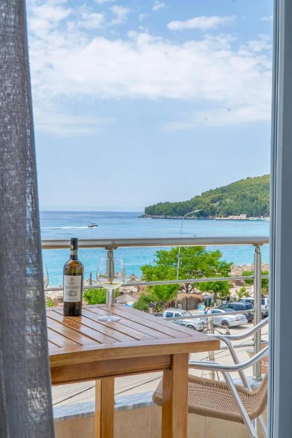 A timber table and a wicker chair on a balcony above the coast road at Himarë, looking across the bay to a pine-covered headland