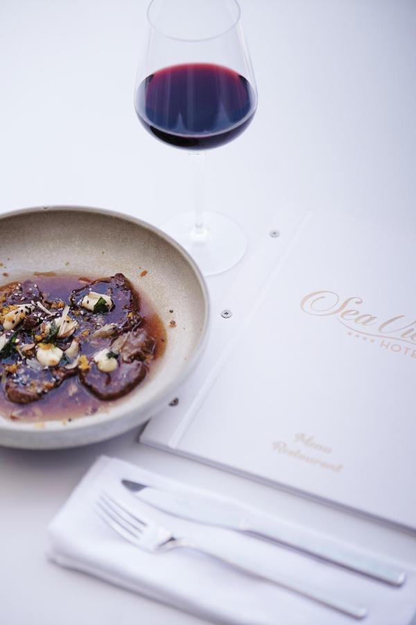 A plated dish of thinly sliced beef in a dark reduction with herbs and shaved cheese, a glass of red wine behind it, and the hotel's white restaurant menu with a gold foil wordmark to the right