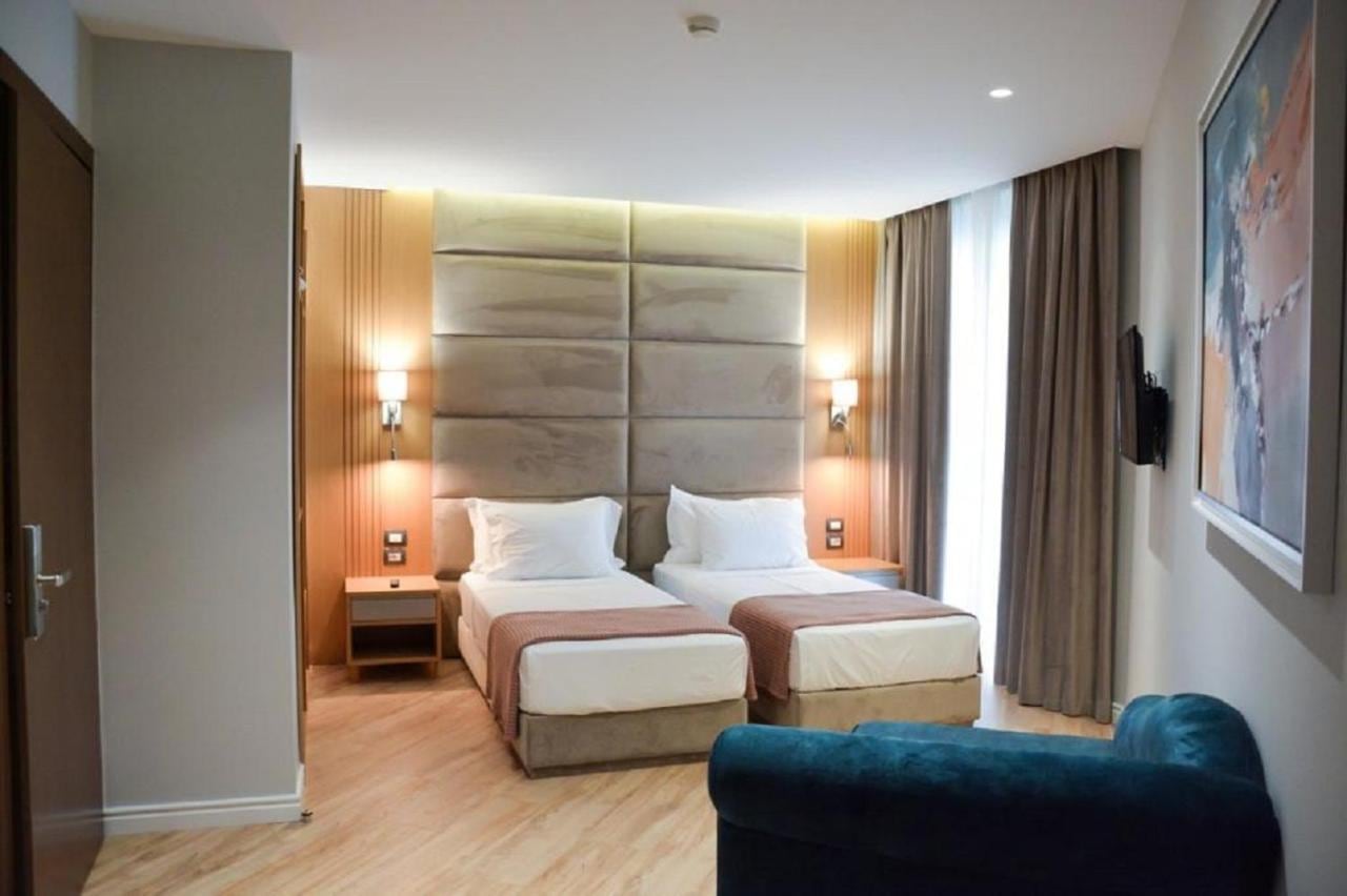 A deluxe twin room with two single beds under a tall padded headboard, timber-lined walls, reading sconces either side, a wall-mounted television and a teal armchair in the foreground