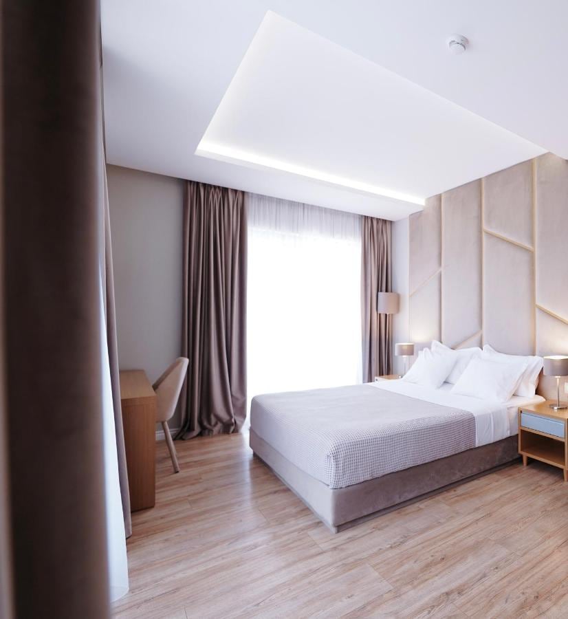 A hotel bedroom with a king bed against a quilted panel headboard, twin bedside lamps, a recessed cove-lit ceiling, a writing desk and chair, and full-length drapes over a balcony door