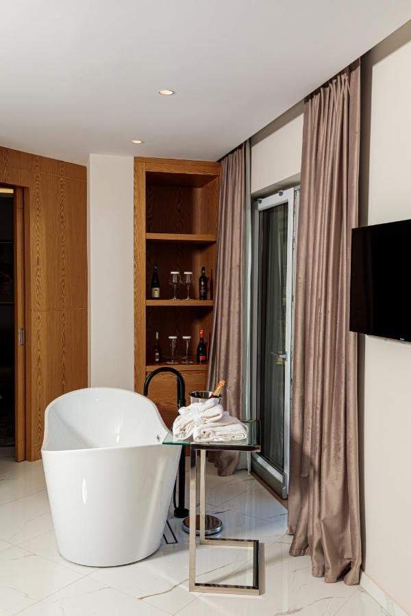 A freestanding white bath with a black floor-standing tap in a suite, beside a glazed balcony door with full-length drapes, a glass side table with folded towels, and a recessed oak shelf holding wine glasses and bottles