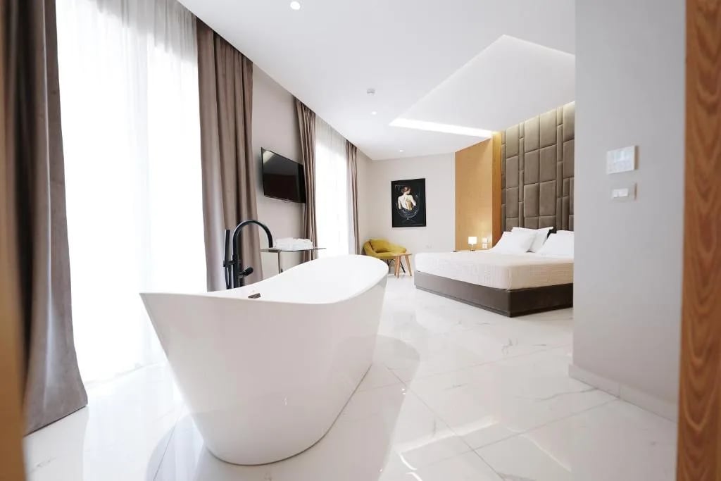 A marble-floored suite with a freestanding white bath in the foreground, a king bed under a quilted headboard beyond, a mustard armchair, a wall-mounted television and full-length drapes