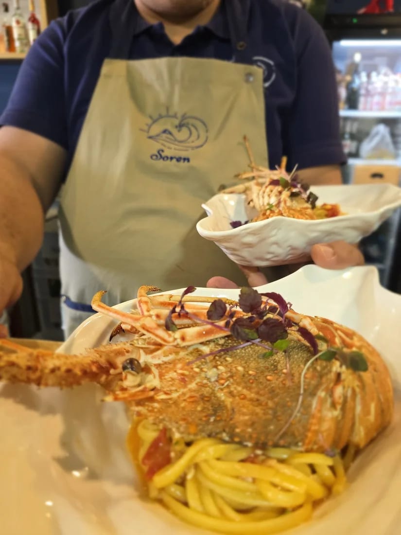 A whole spiny lobster set over linguine on a white dish, carried out by a member of staff in a Soren apron