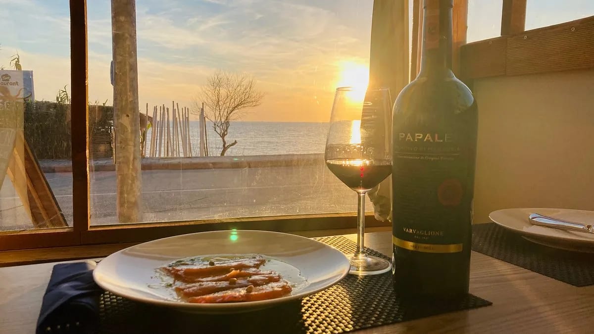 A dish of thinly sliced fish with capers and green oil beside a glass and a bottle of Primitivo, on a table at the window as the sun sets over the Ionian