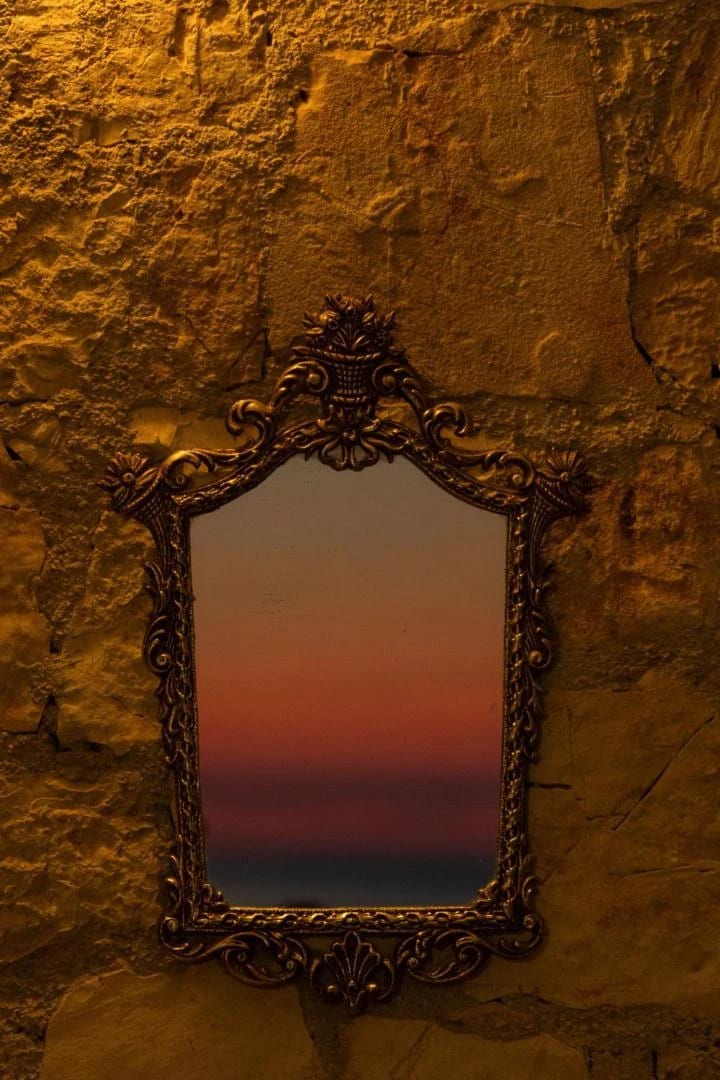 An ornate gilt-framed mirror hung on a stone wall, holding the reflection of the sunset