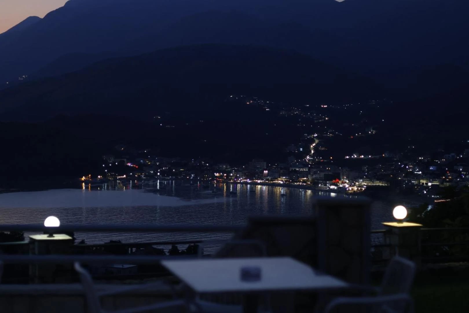 The terrace after dark, lamps lit on the stone wall and the lights of Himarë along the bay below