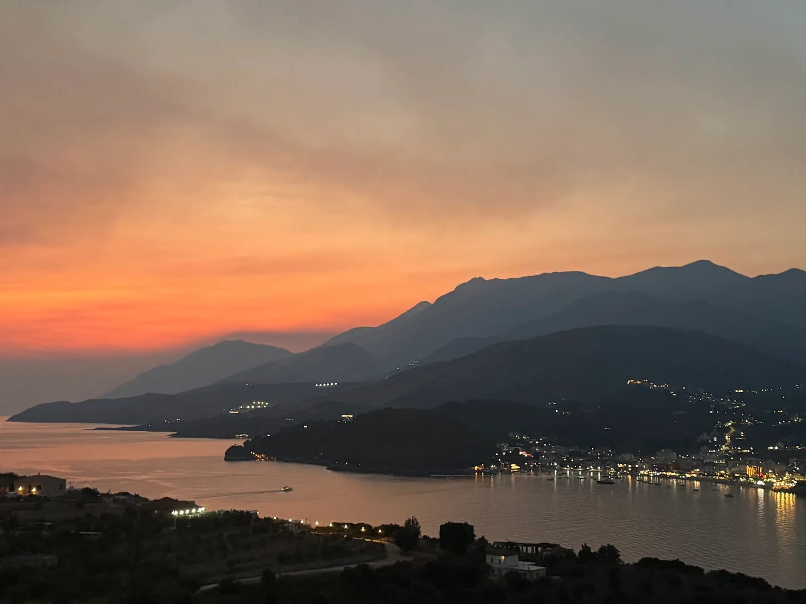 Himarë bay from the terrace just after sunset, orange banding the sky over dark headlands and the town lit along the shore