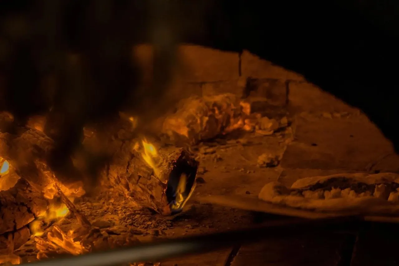 A pizza on the floor of the wood oven beside the burning logs, the brick dome lit orange