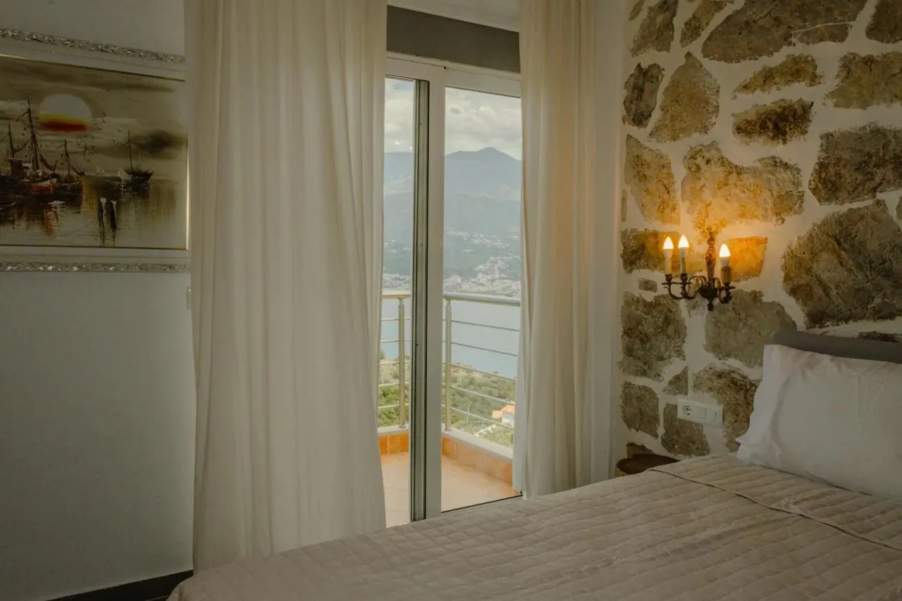 A double bed against a stone wall beside a sliding door onto a balcony, the bay and the headland visible beyond