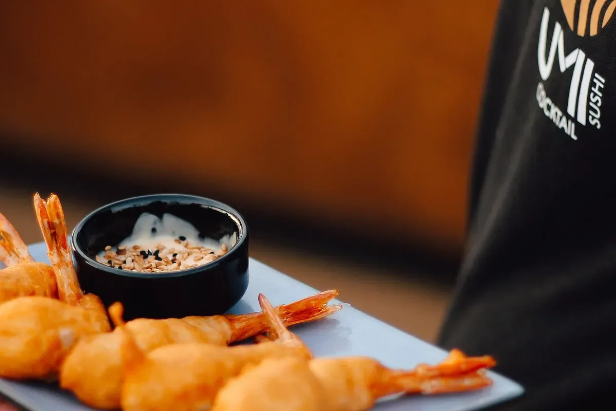 A slate-blue plate of prawns fried in batter with a sesame-topped dip, carried past a black apron printed with UMI's gold fish mark