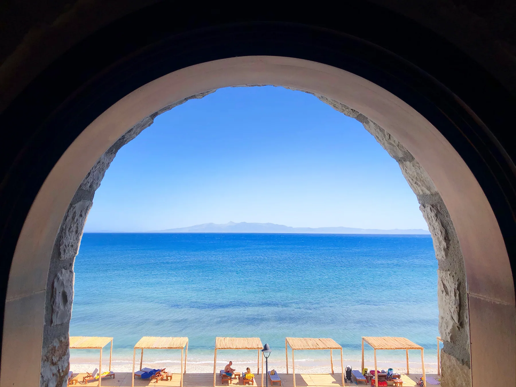 A round stone arch opening straight onto the bay, with the hotel's thatched parasols in a line on the shingle below