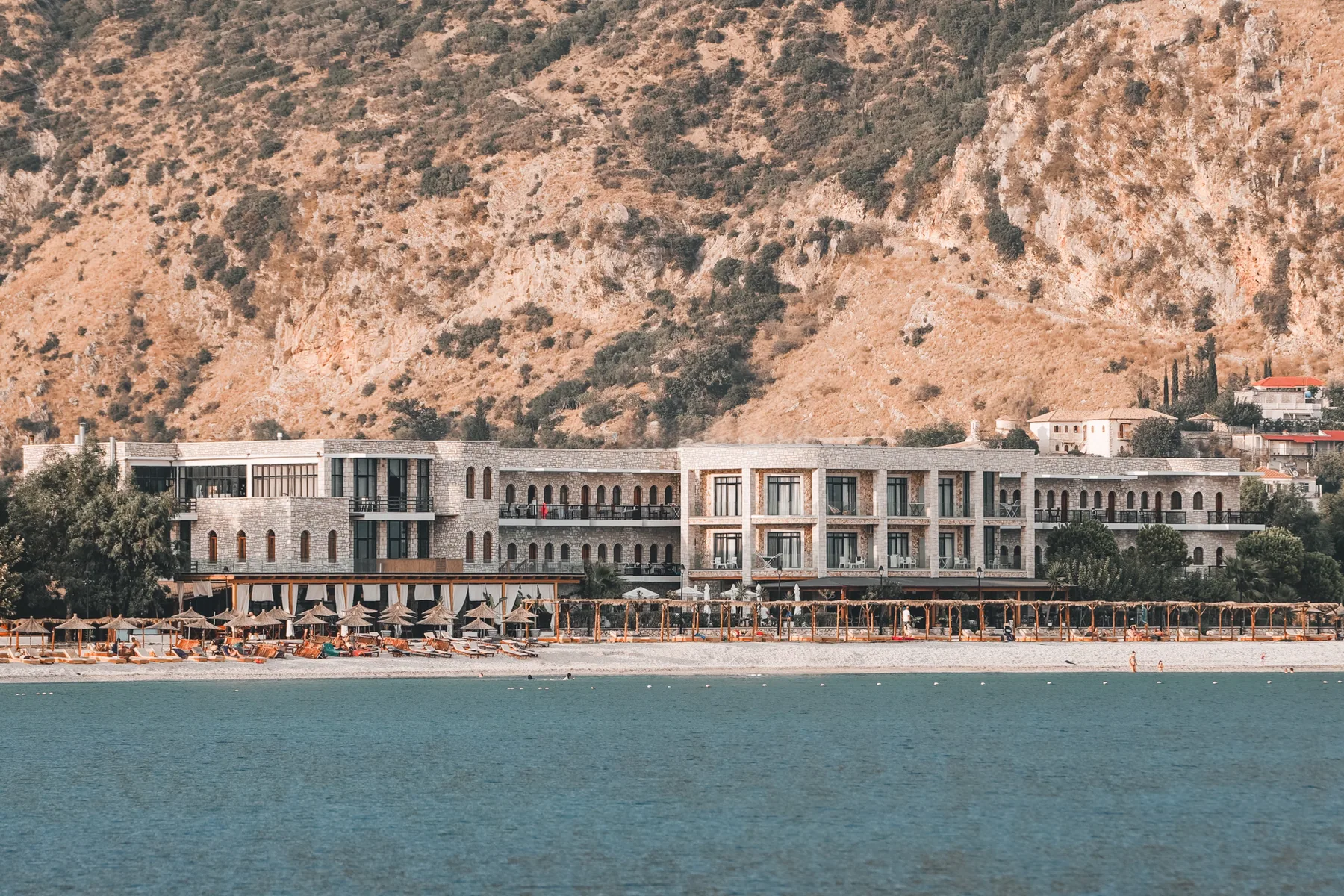 The long limestone front of Valta Hotel photographed from the water, its arched windows in rows, thatched parasols on the shingle below and a bare mountain behind