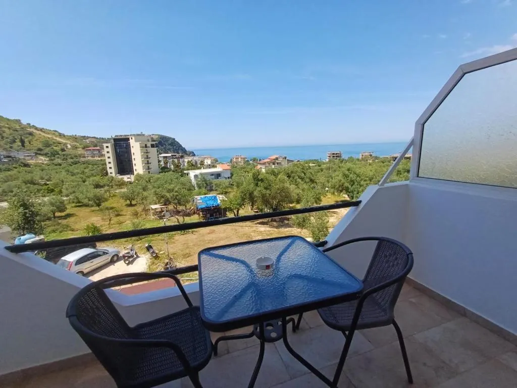 A table and two chairs on a white balcony at Velco BB Himara, looking over an olive grove to the sea and a wooded headland
