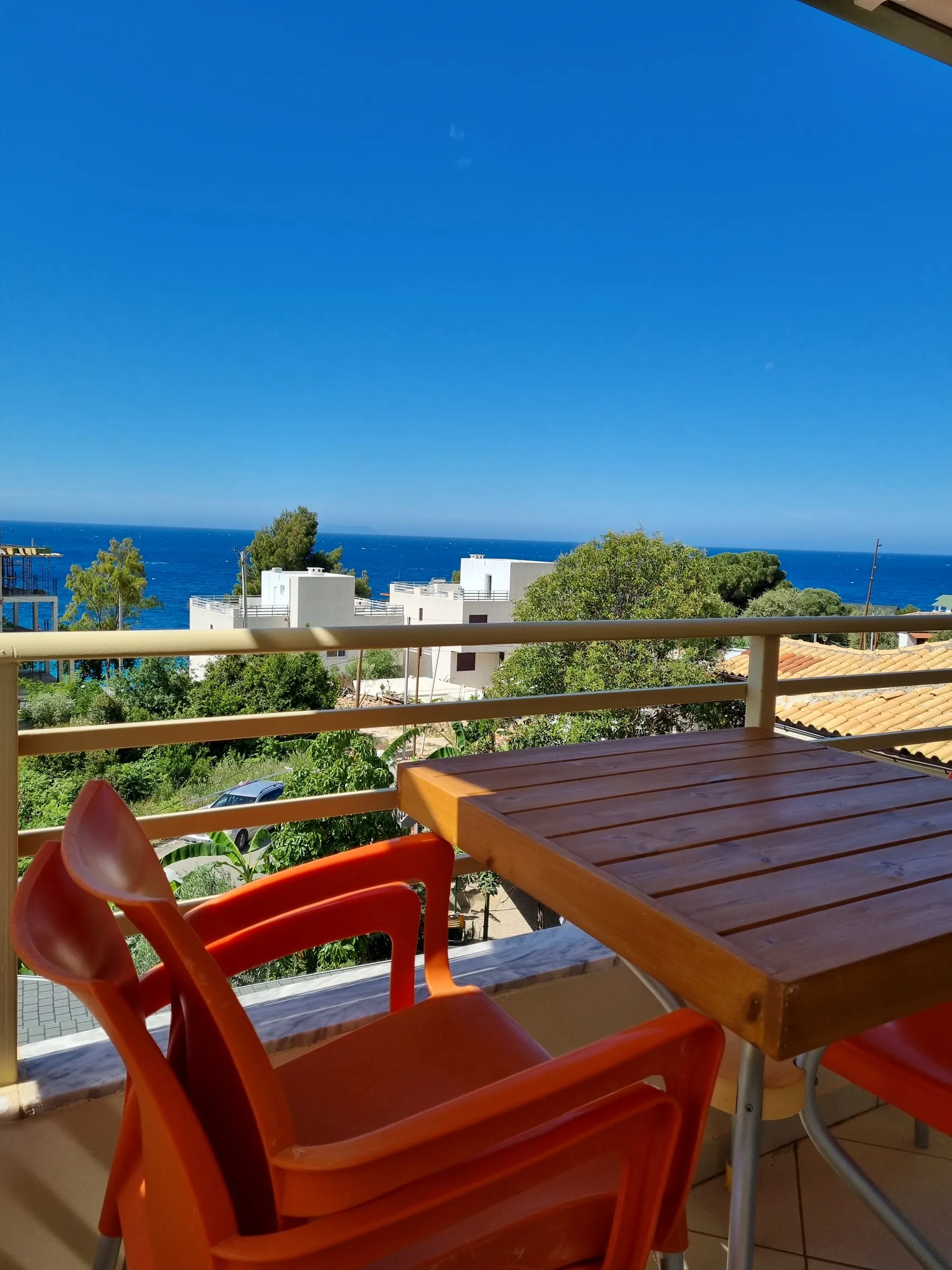 Orange chairs and a timber table on a balcony at Azure Hotel Dhermi, with olive trees, white houses and the sea beyond