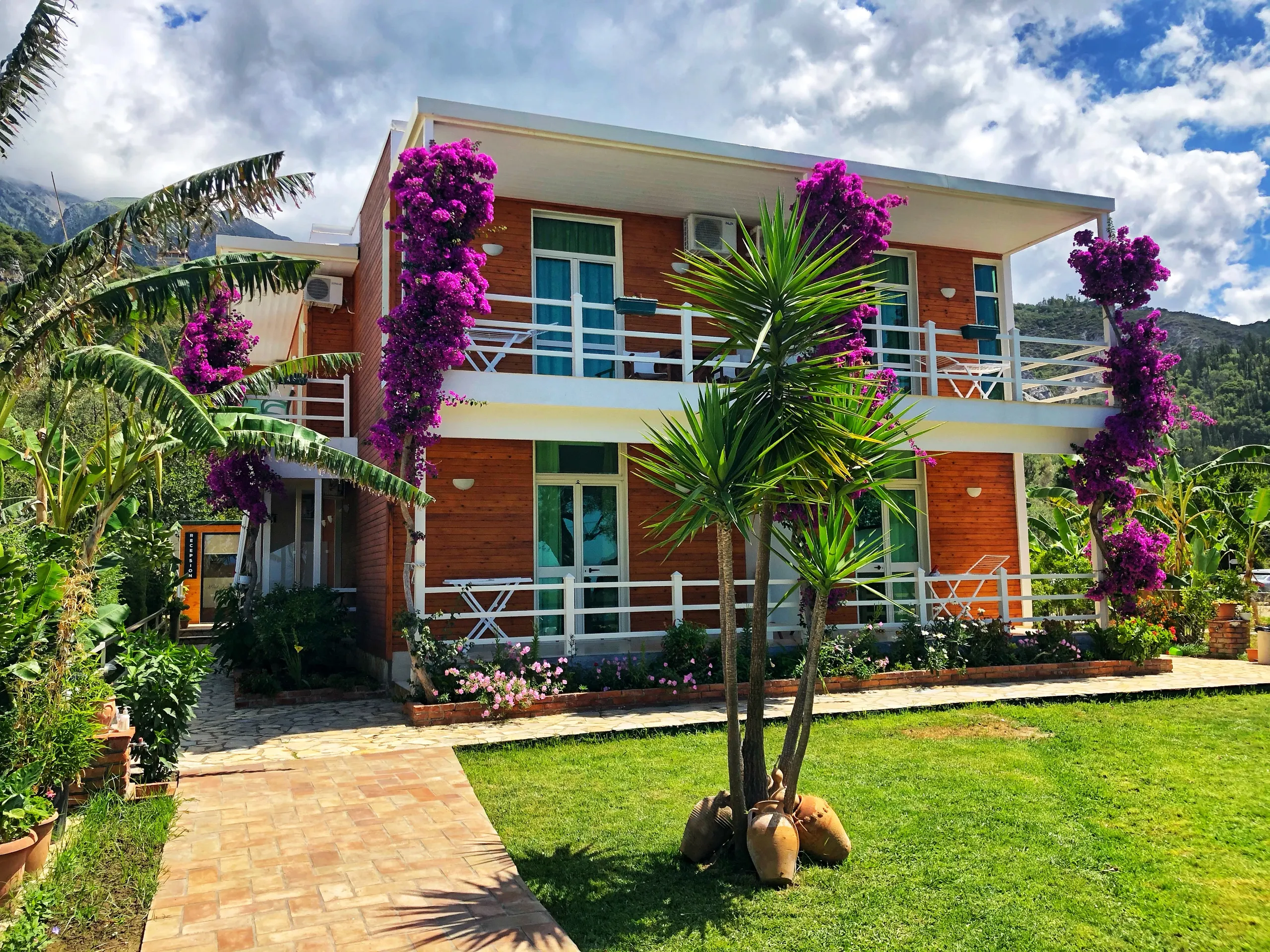 Hotel Jon in Dhërmi: a two-storey house clad in orange-stained timber with white rails and columns of magenta bougainvillea behind its lawn