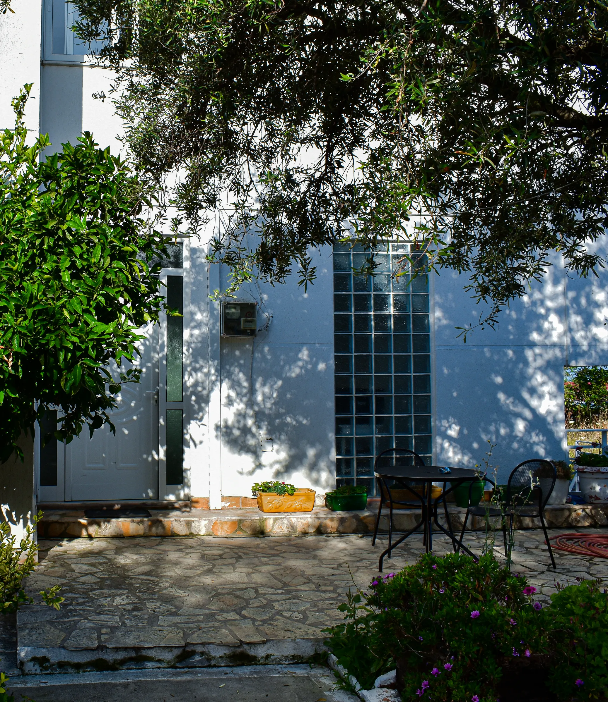 The white front of Hotel Stolis in Potam, Himara, in dappled olive shade, with a glass-block stair window and flagstone steps