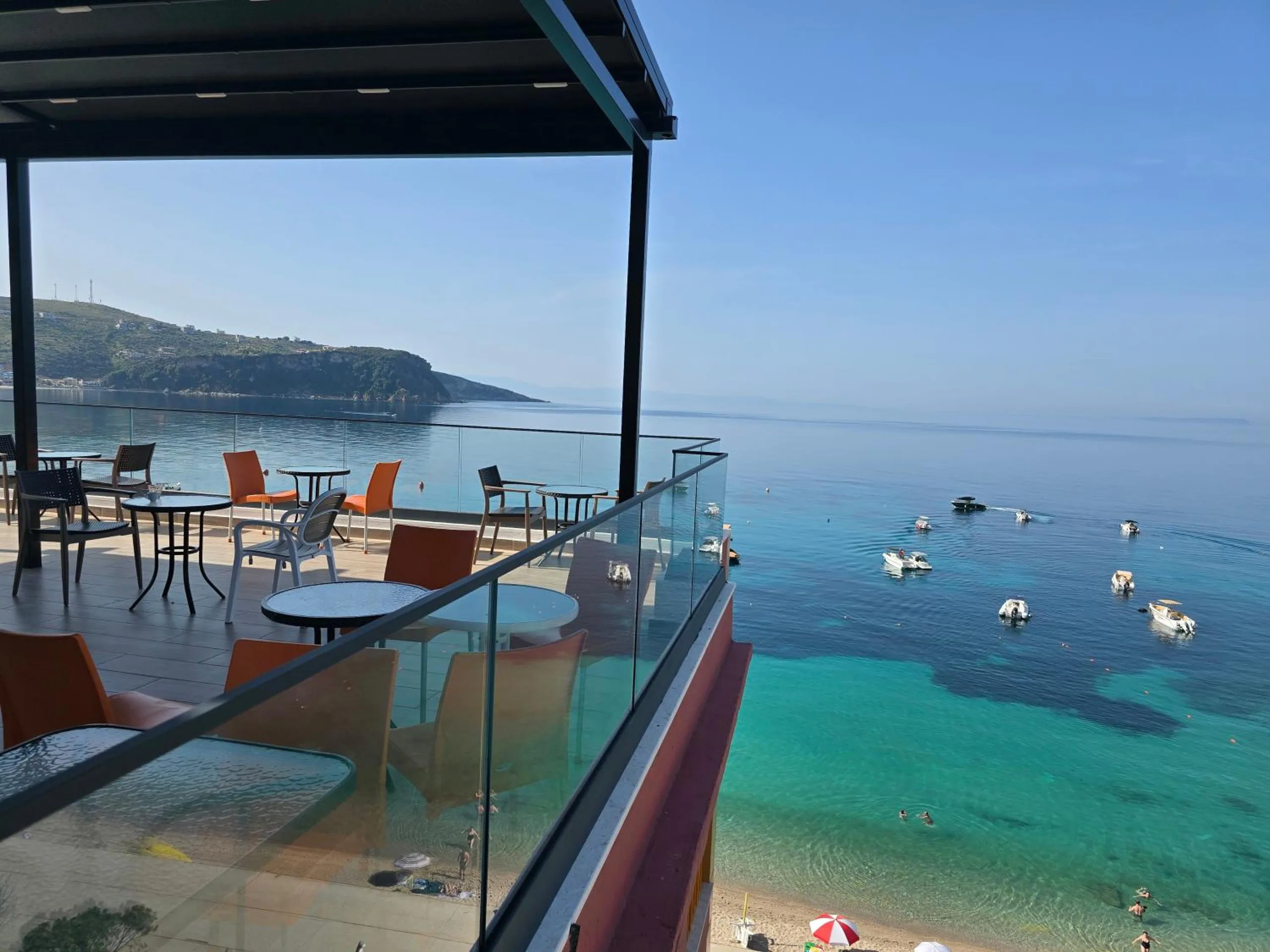 The roof terrace at Hotel Veizi in Himarë, orange chairs behind a glass rail over the turquoise water of Spile bay