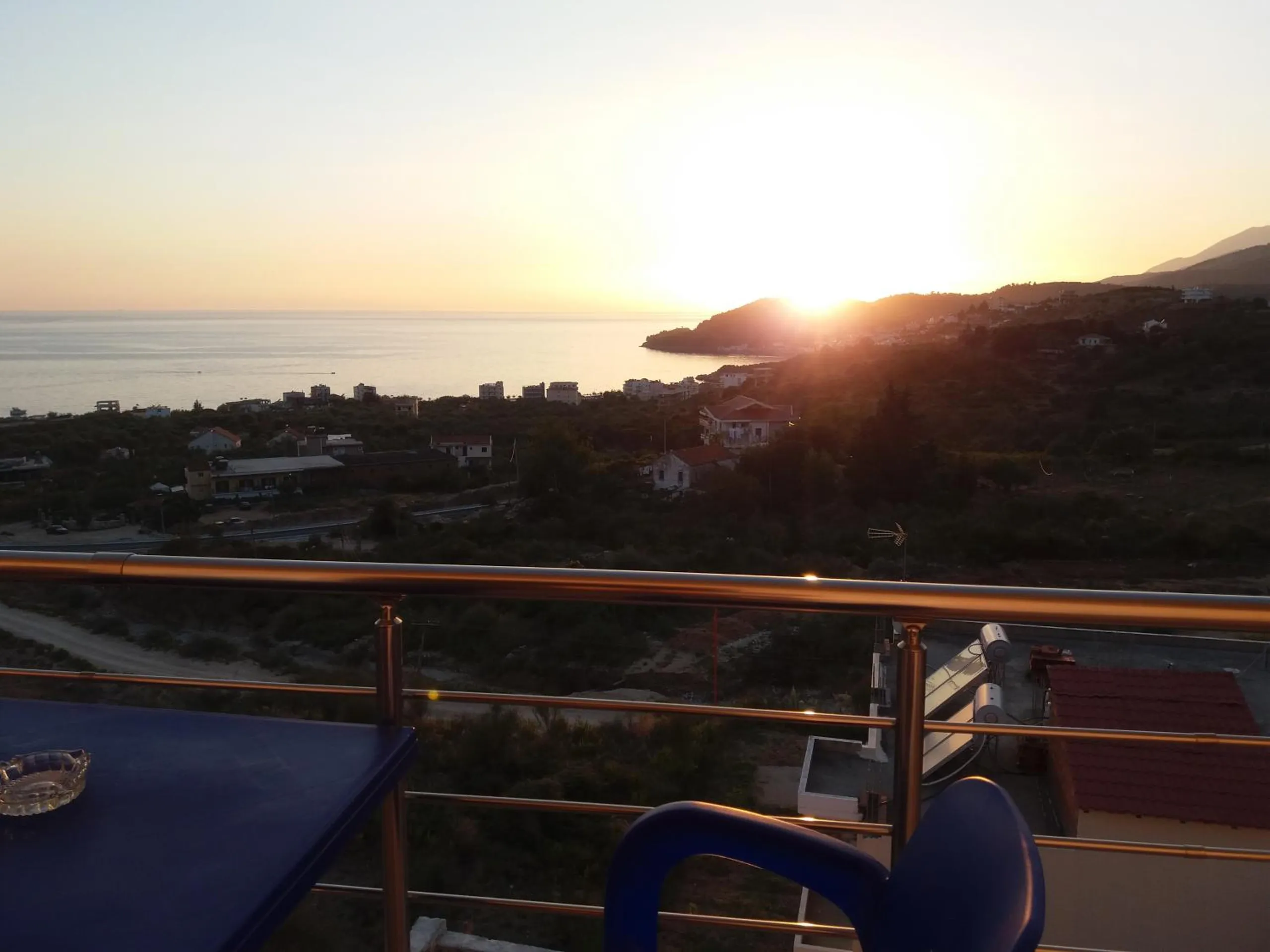 Marika's Apartments in Himara: the sun going down over the bay past a balcony rail and a blue table