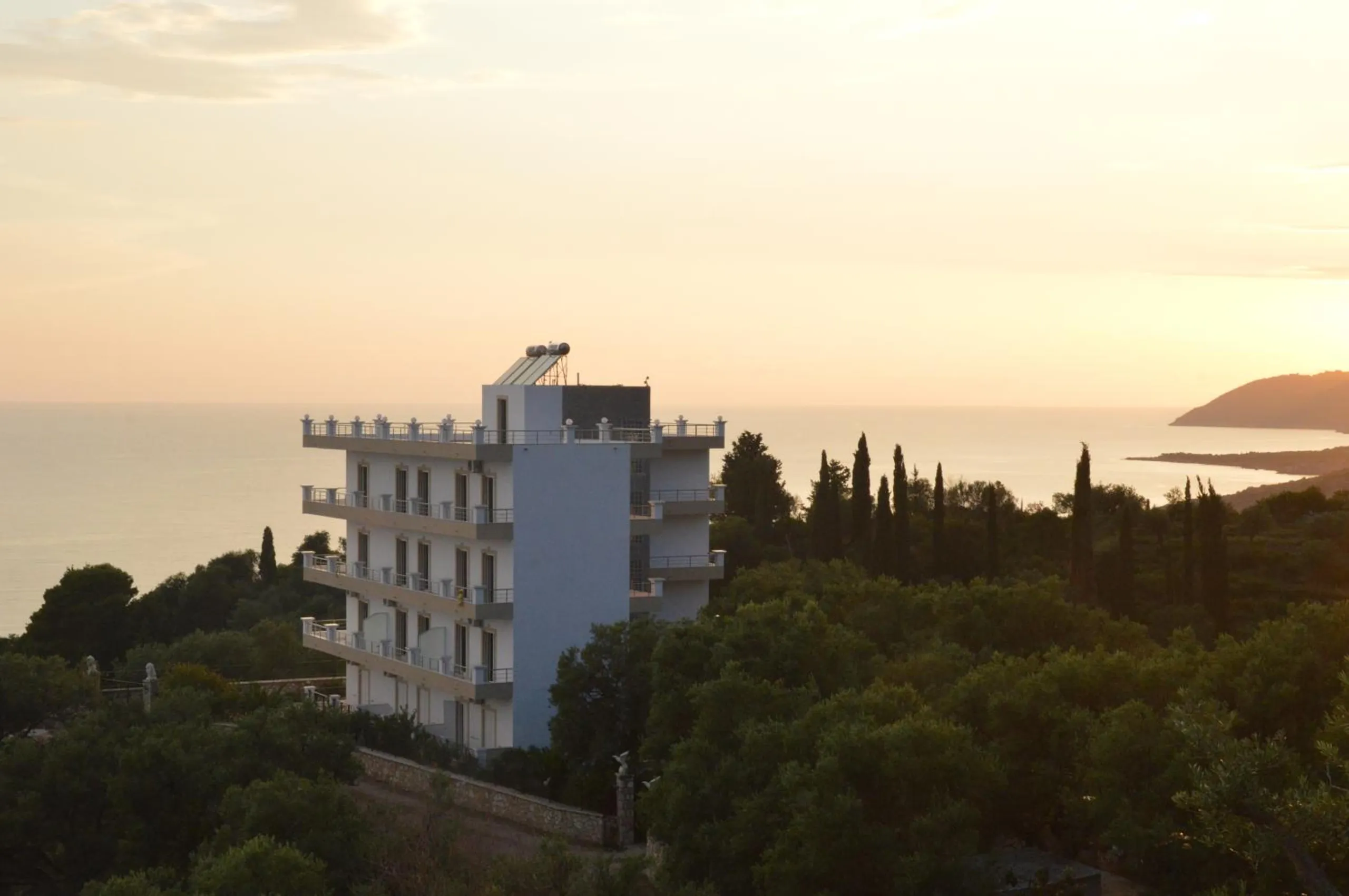 Mihalis Apartments in Himara at sundown: a white five-level house among olive trees and cypresses above the Ionian