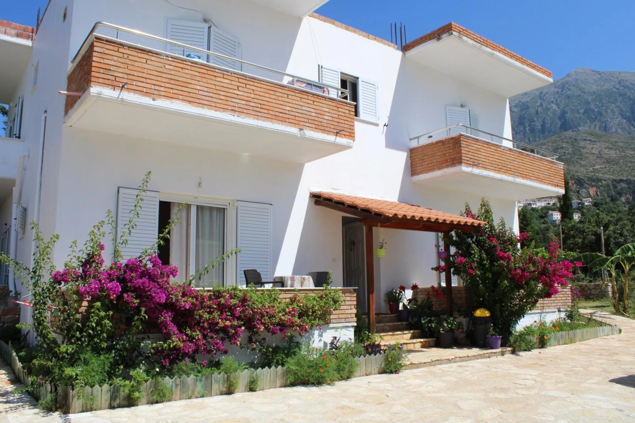 Penelope Rooms in Dhërmi: the white house with brick-faced balconies and magenta bougainvillea along the porch, mountains behind