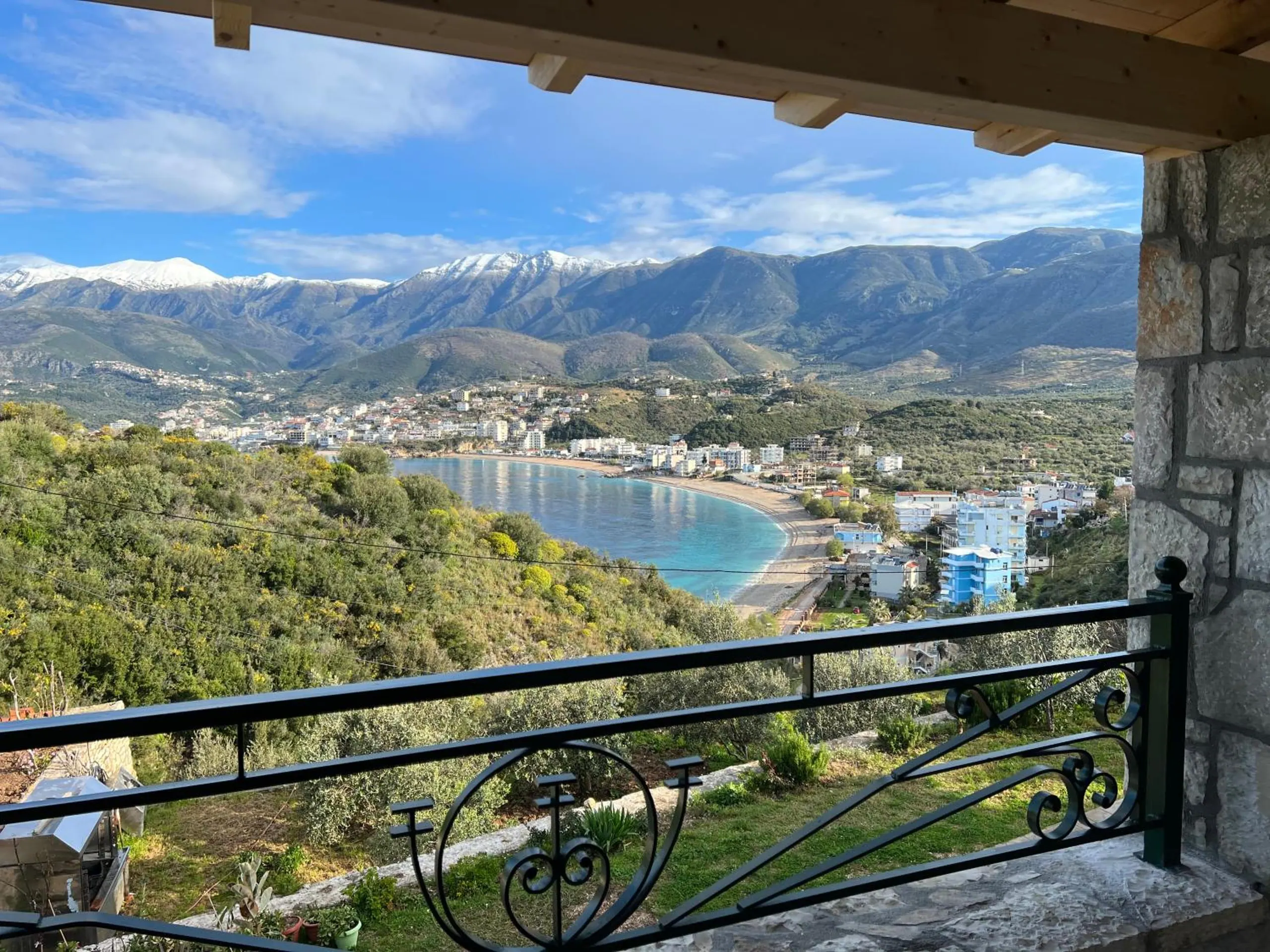 Potam bay from a stone-pillared balcony at Stonevilla Buta in Himara, an iron rail in front and snow on the mountains beyond